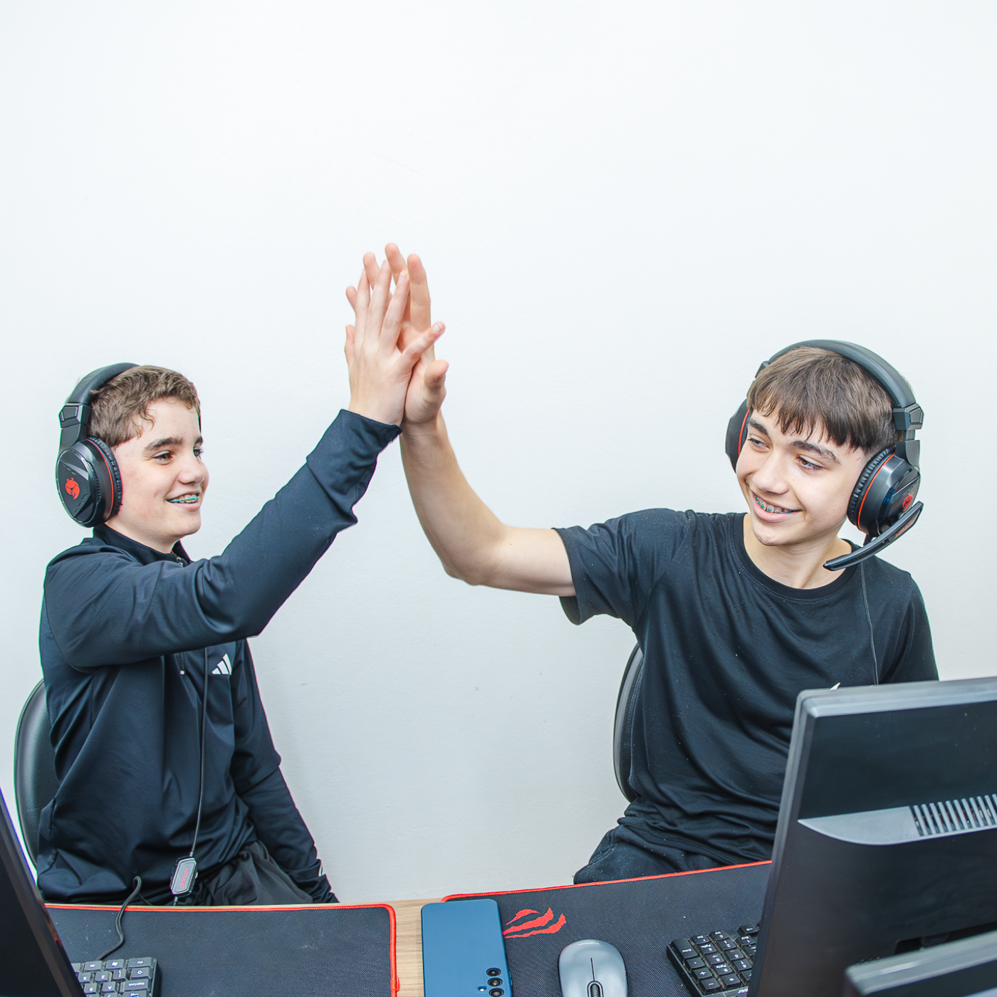 dois meninos usando camiseta preta sentados na frente do computador lado a lado batendo as mãos e sorrindo.