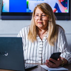 Mulher loira de cabelo comprido e pele branca usando óculos e uma camisa branca com listras pretas sentada na frente do computador e segurando o celular.