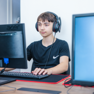 jovem de cabelo escuro e pele clara sentado na frente do computador usando fones de ouvido pretos e usando uma camiseta preta