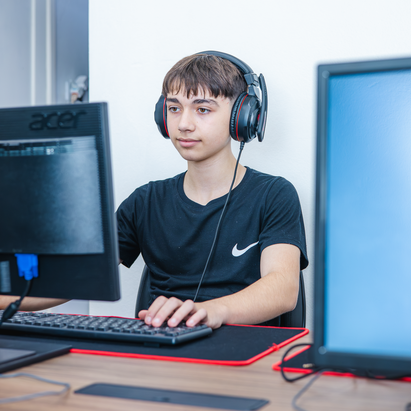 jovem de cabelo escuro e pele clara sentado na frente do computador usando fones de ouvido pretos e usando uma camiseta preta